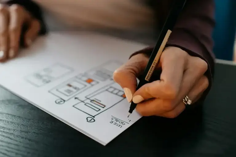 hands-drawing-a-website-wireframe-on-paper-1webp | Lobo Digital | Lobo Digital Close-up of a hand drawing a diagram on white paper with a black pencil, showing boxes and icons like a schematic layout on a dark surface.